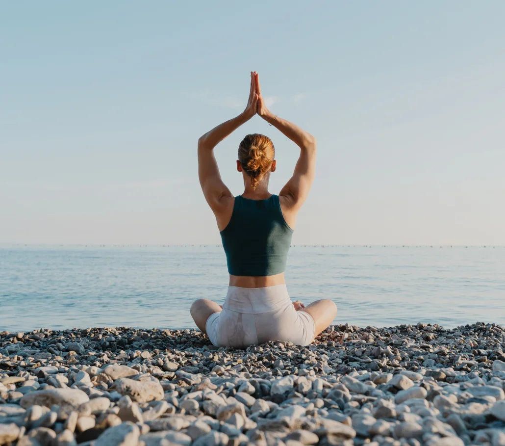 Woman practicing yoga in a seated meditation pose on a rocky beach by the ocean.