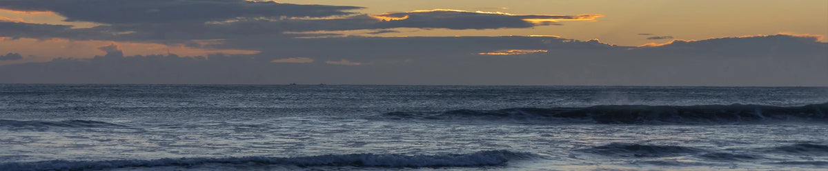 A wide panoramic view of ocean waves under a dramatic sunset sky with dark clouds.