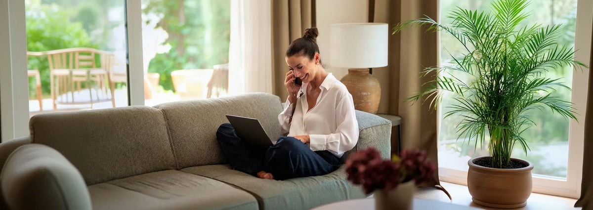 Smiling woman sitting on a sofa using a laptop and talking on a phone in a bright living room.